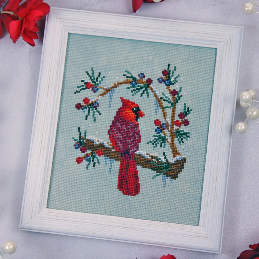 A framed cross-stitch artwork featuring a red cardinal perched on a snowy branch with blue berries, set in a white frame on a light background with decorative red flowers and pearl beads.
