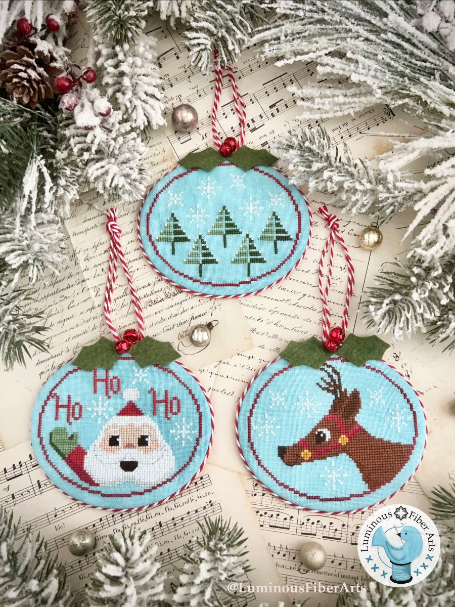 Cross-stitched Christmas ornaments with Santa, reindeer, and 'Ho Ho' text on a textured surface with pine branches.