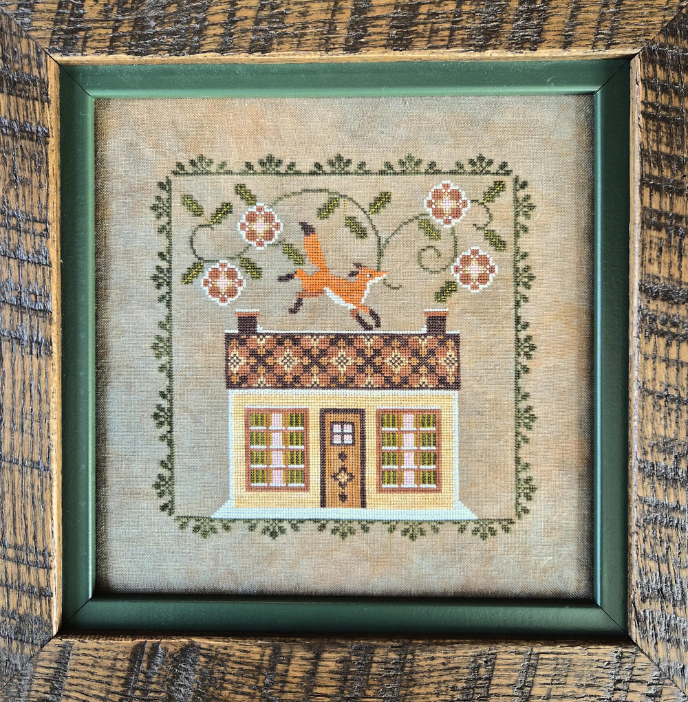 A framed needlepoint or embroidery artwork showing a small house with a patterned roof, floral vine border, and a fox jumping over the roof. The piece is centered in a dark green frame against a rustic wood background.