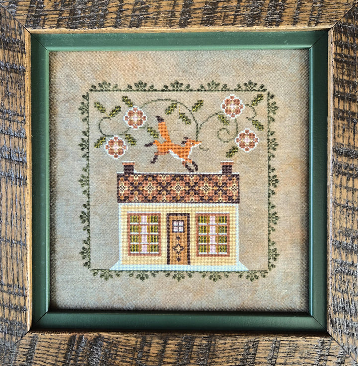 A framed needlepoint or embroidery artwork showing a small house with a patterned roof, floral vine border, and a fox jumping over the roof. The piece is centered in a dark green frame against a rustic wood background.
