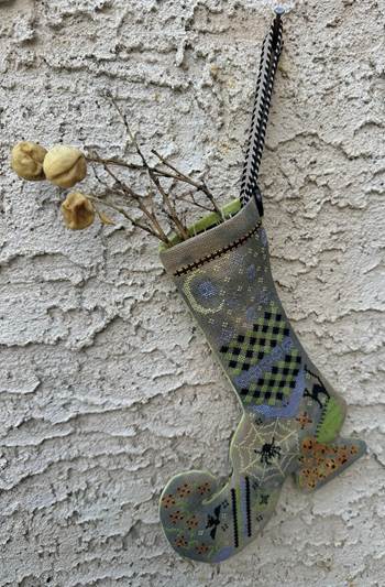 A handmade patchwork Christmas stocking hanging on a textured stucco wall, filled with dried branches and small yellow decorative buds. The stocking features various patterned panels including black and green checkered squares, a spiderweb motif, and a small orange patterned section.