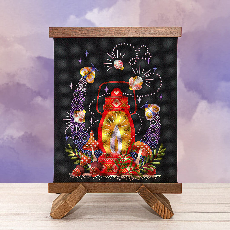 A vertical black needlepoint canvas displayed on a small wooden easel stand, featuring a colorful cross-stitch design of a lantern, mushrooms, flowers, and fireworks against a purple cloudy sky backdrop.