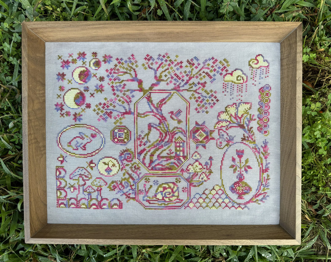 A framed cross-stitch artwork displayed outdoors on green grass. The piece features a whimsical, folk-art style design with a tree, moon phases, clouds, flowers, and animals, stitched in pink, purple, yellow, and green threads on white fabric, enclosed in a light brown wooden frame.