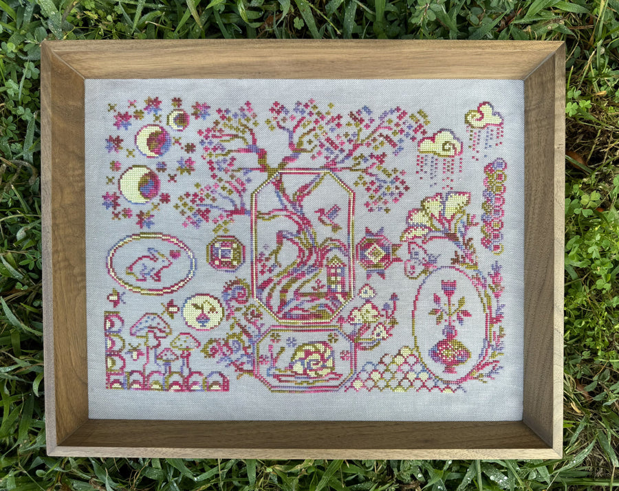 A framed cross-stitch artwork displayed outdoors on green grass. The piece features a whimsical, folk-art style design with a tree, moon phases, clouds, flowers, and animals, stitched in pink, purple, yellow, and green threads on white fabric, enclosed in a light brown wooden frame.