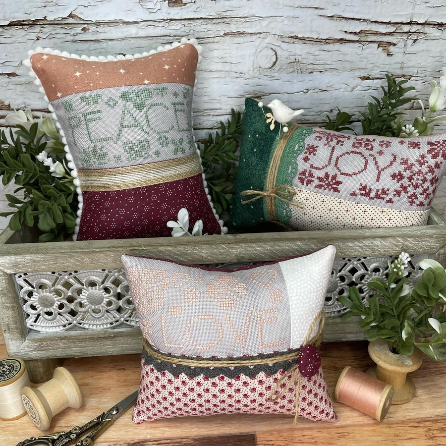 Decorative pillows with 'Peace', 'Joy', and 'Love' text on a rustic wooden surface with greenery.