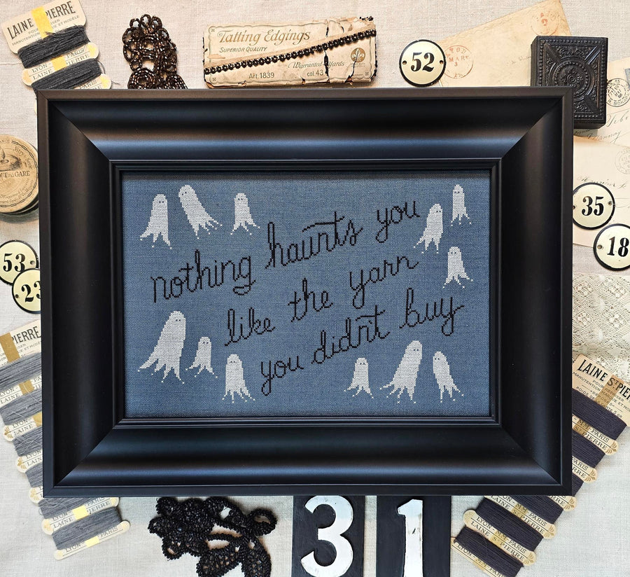 A framed cross-stitch piece with a black frame, featuring white ghost illustrations and the phrase 'nothing haunts you like the yarn you didn't buy' on a blue-gray fabric background, surrounded by vintage-style yarn packaging and decorative props.