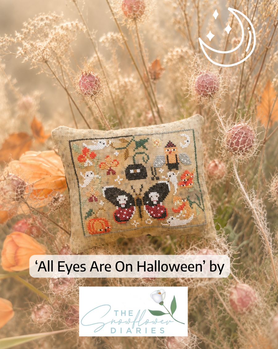 A square embroidered pillow displayed in a field of dried flowers. The pillow features a cross-stitch design of a black and red butterfly with pumpkins, ghosts, and a haunted house, with the text 'All Eyes Are On Halloween' by The Smoother Diaries.