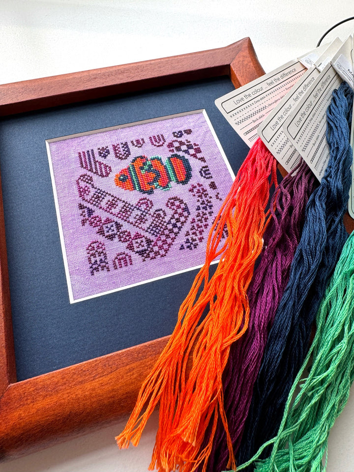 Framed cross-stitched picture of a clownfish on a purple background with colorful embroidery floss beside it.