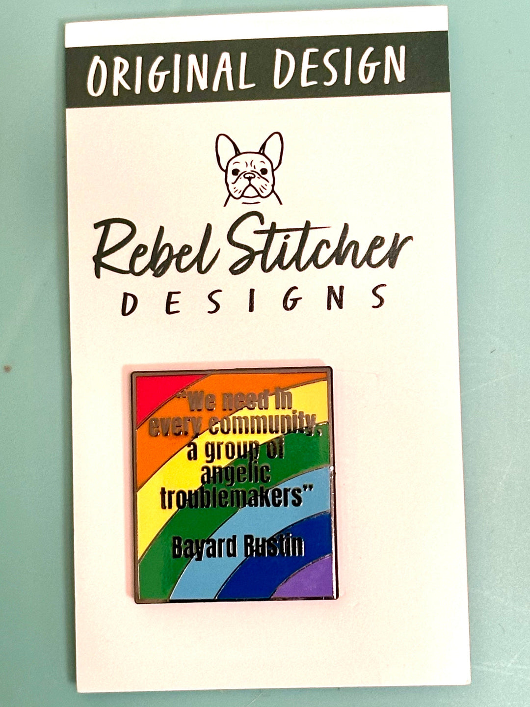 Pin with rainbow design and text on a card labeled 'Rebel Stitcher Designs' against a light green background