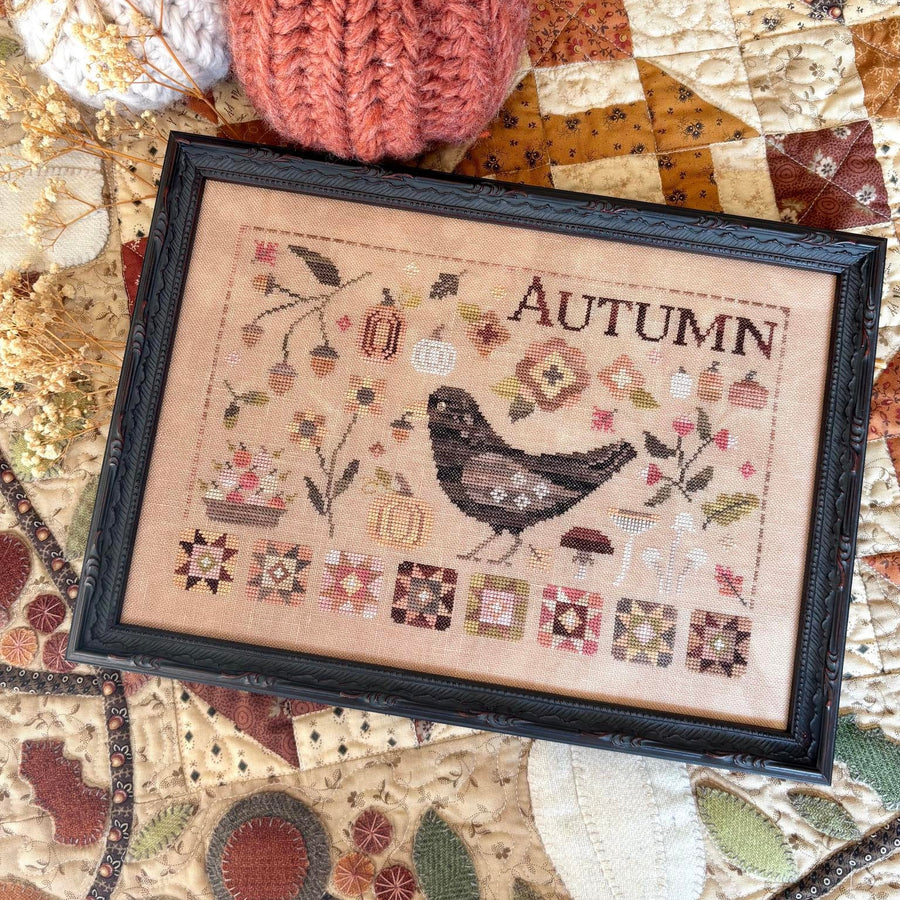 A framed autumn-themed cross-stitch piece with a bird and the word 'AUTUMN,' styled on a quilted fabric surface with decorative pumpkins nearby.