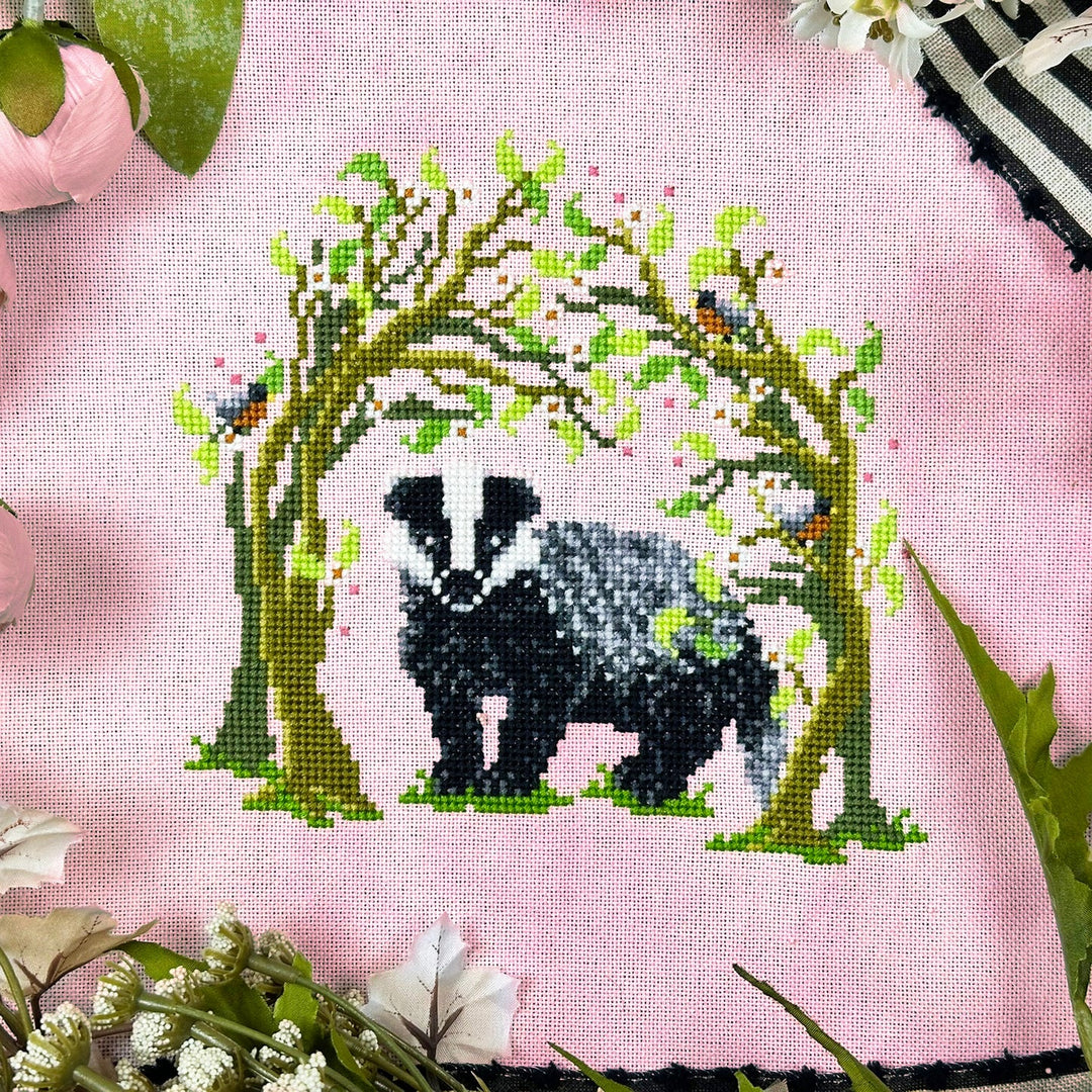 Cross-stitched design of a badger in a floral arch on pink fabric.