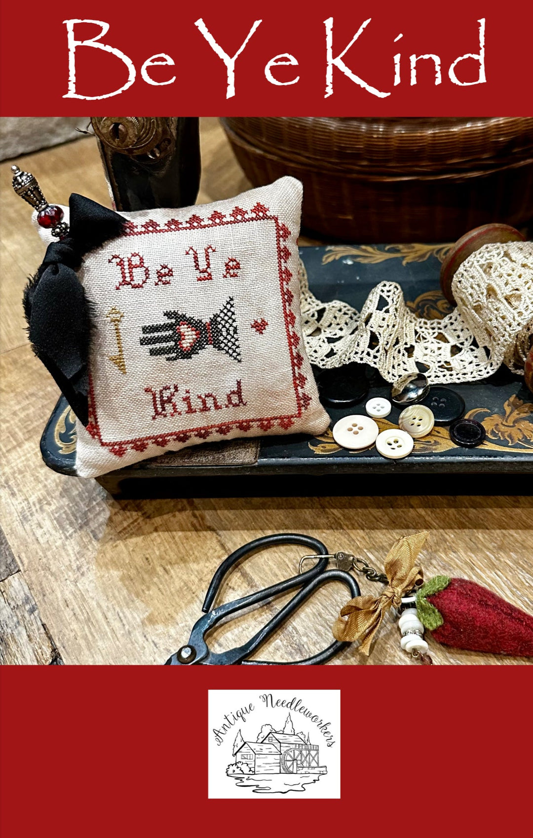 Decorative pillow with cross-stitch design and text 'Be Ye Kind' on a wooden surface with sewing supplies.