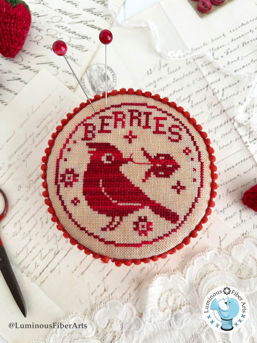 Cross-stitched design of a cardinal with berries on fabric, pinned to a surface.