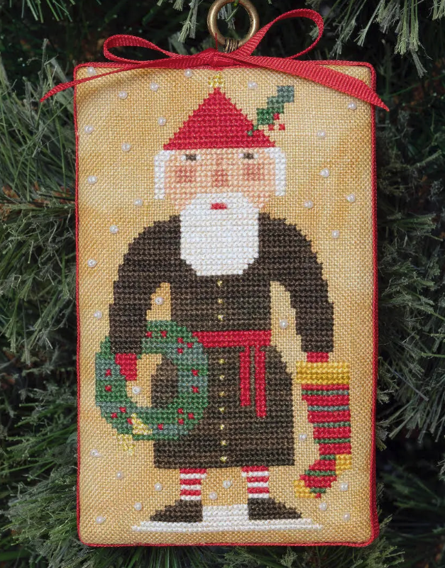 Cross-stitched ornament of a traditional Santa Claus figure with a wreath, hanging on a tree.