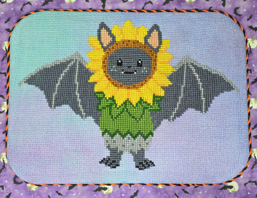 A square cross-stitch or needlepoint canvas featuring a cute cartoon bat wearing a sunflower head and green dress, bordered by an orange and black stitched edge on a purple patterned fabric background.