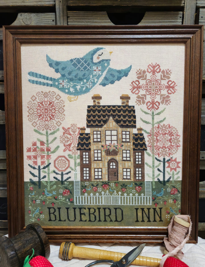 Framed cross-stitched artwork of 'Bluebird Inn' with a house, flowers, and a blue bird.