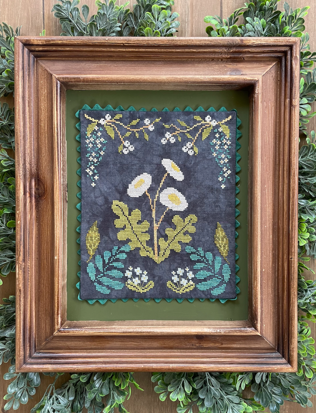 A framed cross-stitch needlework depicting floral designs, displayed within a wooden frame, set against a background of green foliage.