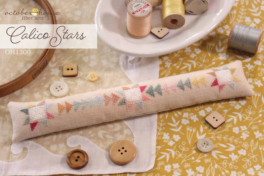 Cross-stitched pillow with 'Calico Stars' design on a fabric background with spools of thread and buttons.