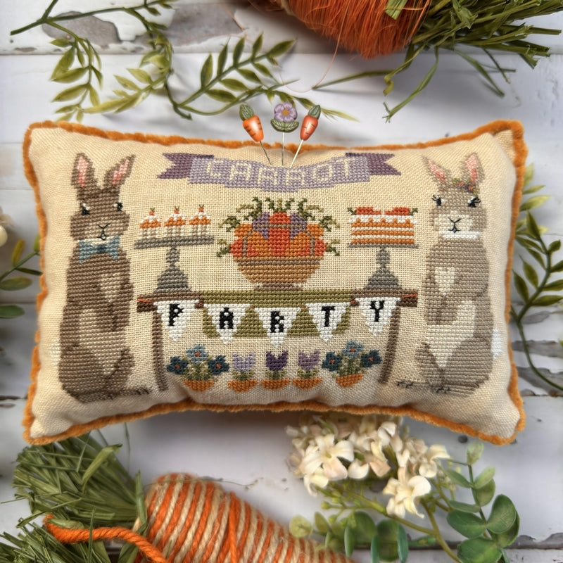 Decorative pillow with cross-stitched design of bunnies and 'PARTY' text, surrounded by greenery.