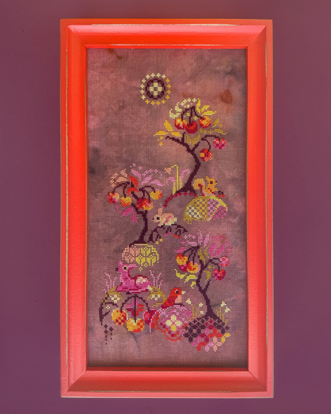 A framed vertical needlepoint or cross-stitch artwork featuring a stylized tree with colorful leaves, birds, and floral motifs on a mauve background, set in a bright coral-red frame against a solid dark purple wall.
