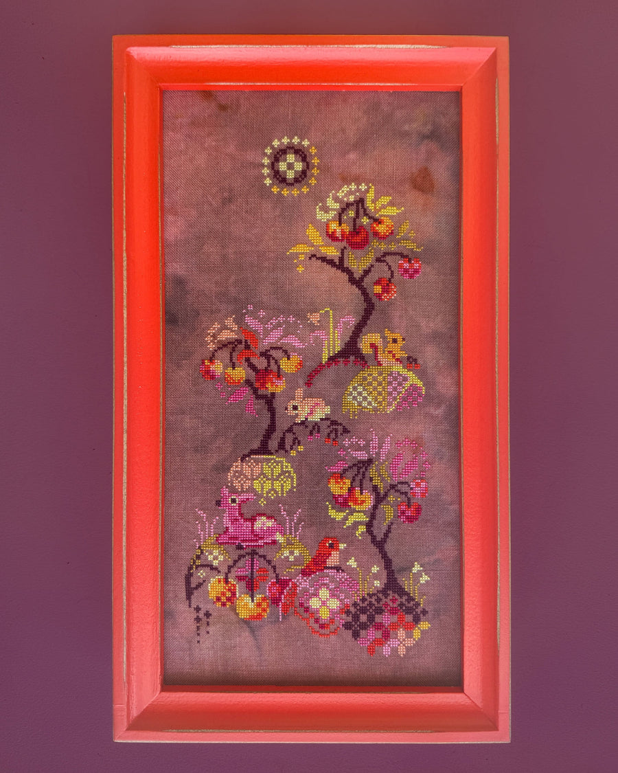 A framed vertical needlepoint or cross-stitch artwork featuring a stylized tree with colorful leaves, birds, and floral motifs on a mauve background, set in a bright coral-red frame against a solid dark purple wall.
