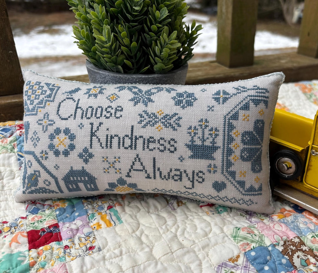 Decorative pillow with cross-stitch design and text 'Choose Kindness Always' on a quilted surface.