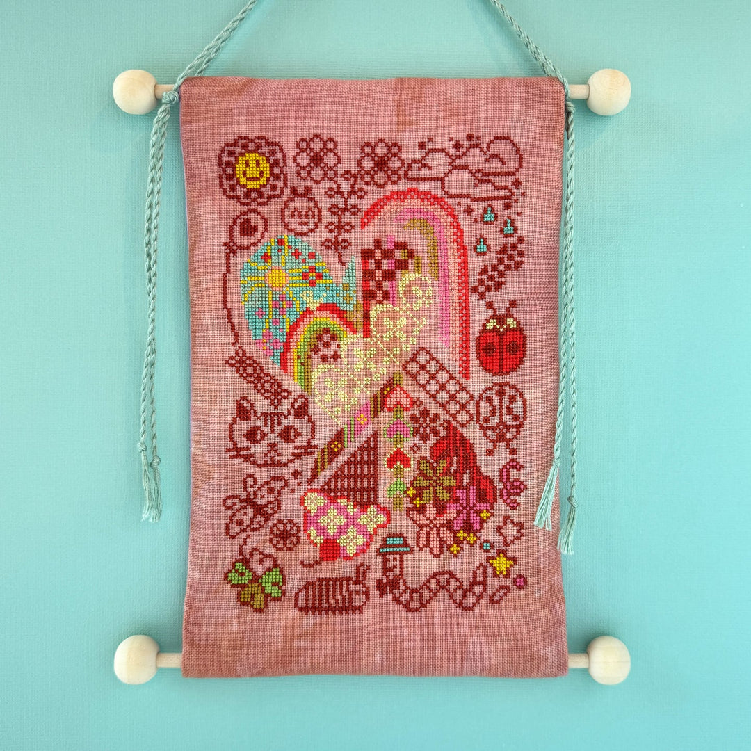 Cross-stitched design on a pink fabric with colorful heart and floral patterns, hanging against a light blue background.
