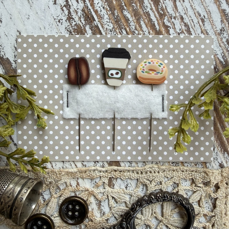 A flat-lay of three miniature coffee-themed pins (two coffee beans and a takeaway coffee cup) on a white felt bar, mounted on a beige and white polka dot backing card. The card is styled on a rustic wooden surface with lace, buttons, and greenery.