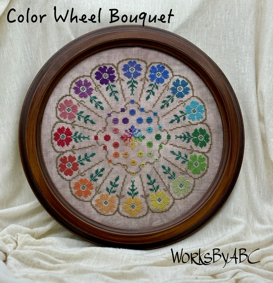 Cross-stitched floral design on fabric with a wooden frame, labeled 'Color Wheel Bouquet' by 'WorksByABC'.