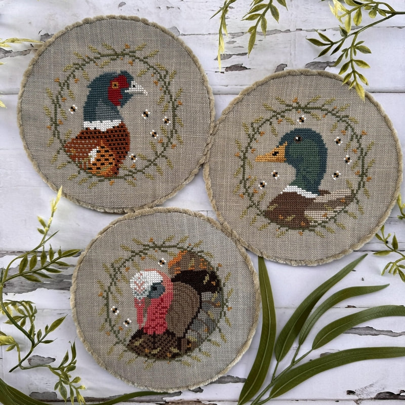 Three needlepoint circles with bird designs on a wooden surface with green leaves.