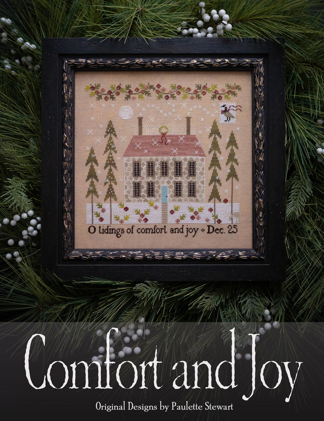 Framed cross-stitched piece with a house design and Christmas text, surrounded by greenery.