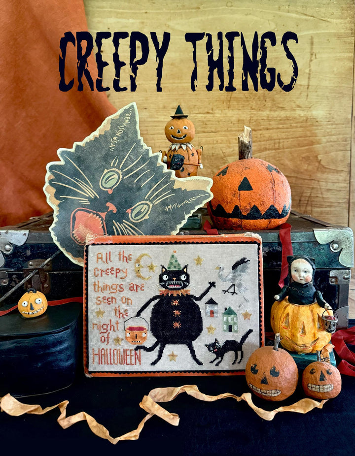 Halloween-themed decor with pumpkins, a cat cross-stitch, and decorative items on a wooden surface.
