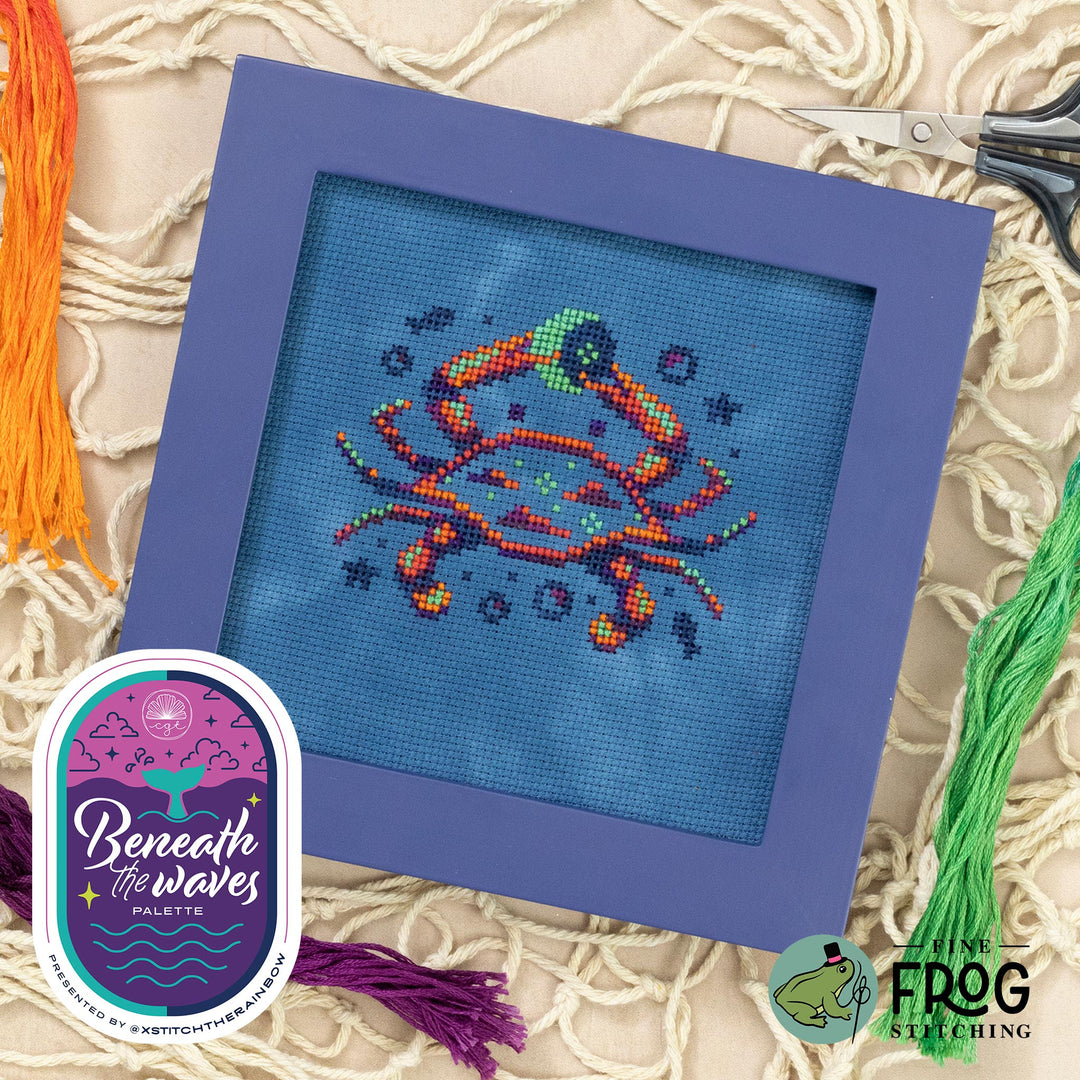 Cross-stitched crab design in a purple frame with embroidery threads and scissors on a textured fabric background.