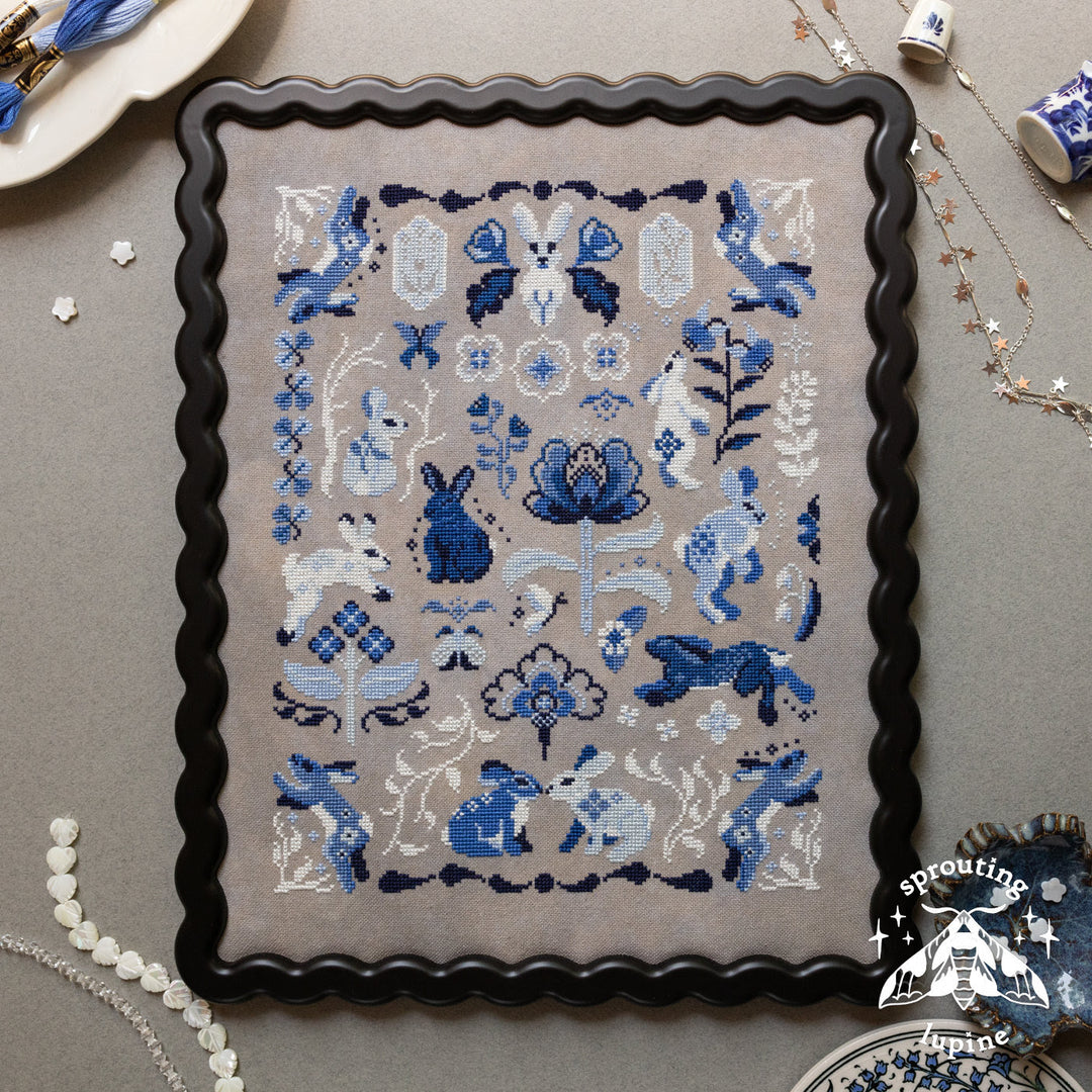 A framed needlepoint canvas featuring a blue and white rabbit and floral design on a beige background, surrounded by a black scalloped frame. The canvas is displayed on a gray surface with decorative items like pearl beads, a ceramic dish, and jewelry.