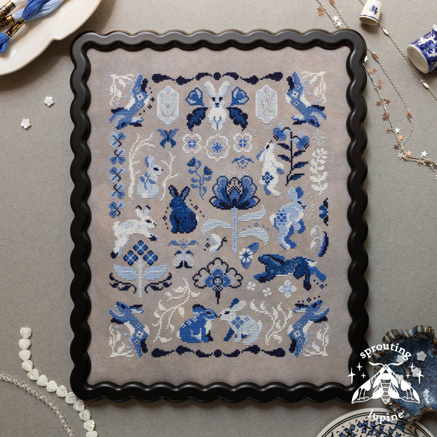 A framed needlepoint canvas featuring a blue and white rabbit and floral design on a beige background, surrounded by a black scalloped frame. The canvas is displayed on a gray surface with decorative items like pearl beads, a ceramic dish, and jewelry.