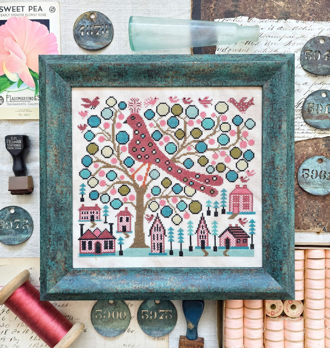 Framed cross-stitch pattern with a bird and houses on a tree, surrounded by vintage items.