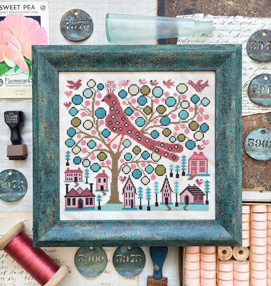 Framed cross-stitch pattern with a bird and houses on a tree, surrounded by vintage items.