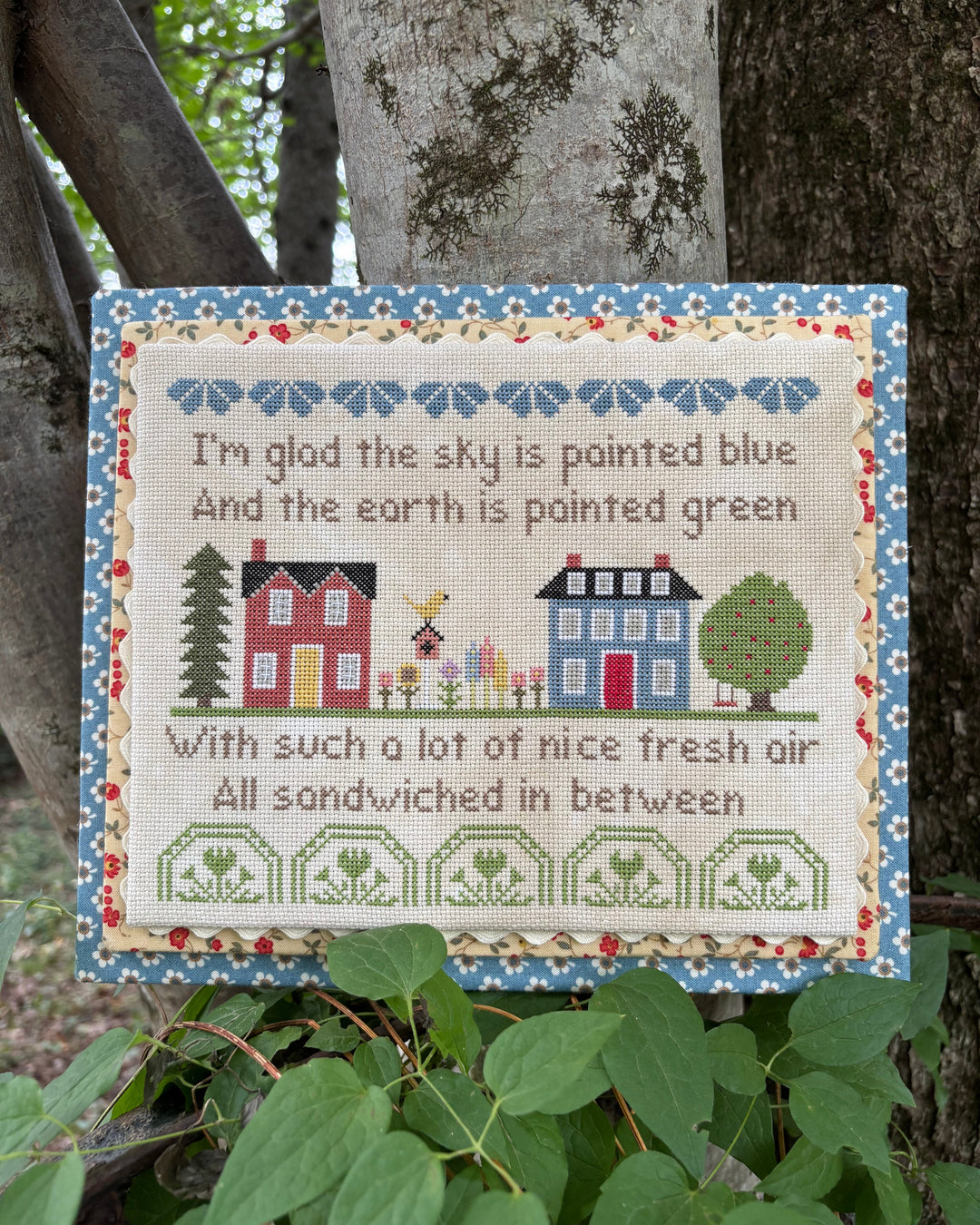 A square embroidered cross-stitch sampler hanging on a tree trunk outdoors. The sampler features a floral blue border, a cream center, embroidered houses, trees, and a bee, with the text: 'I'm glad the sky is painted blue And the earth is painted green With such a lot of nice fresh air All sandwiched in between.