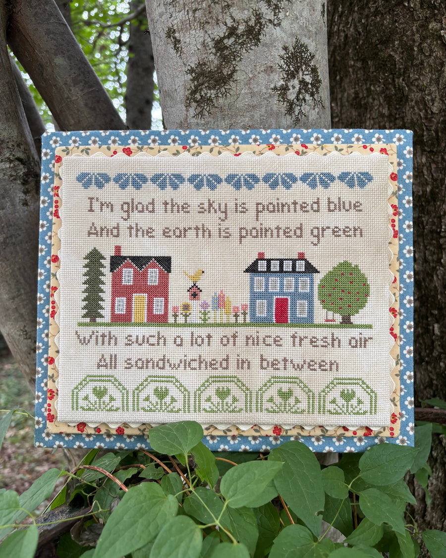 A square embroidered cross-stitch sampler hanging on a tree trunk outdoors. The sampler features a floral blue border, a cream center, embroidered houses, trees, and a bee, with the text: 'I'm glad the sky is painted blue And the earth is painted green With such a lot of nice fresh air All sandwiched in between.