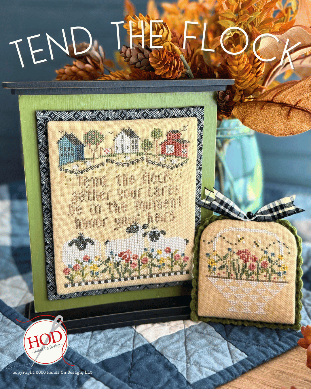A cross-stitch pattern display titled 'TEND THE FLOCK' showing a framed finished piece with a barn, sheep, and floral border, alongside a small matching embroidered pouch. The scene includes decorative autumn dried flowers, a quilted surface, and a visible HOD logo watermark.