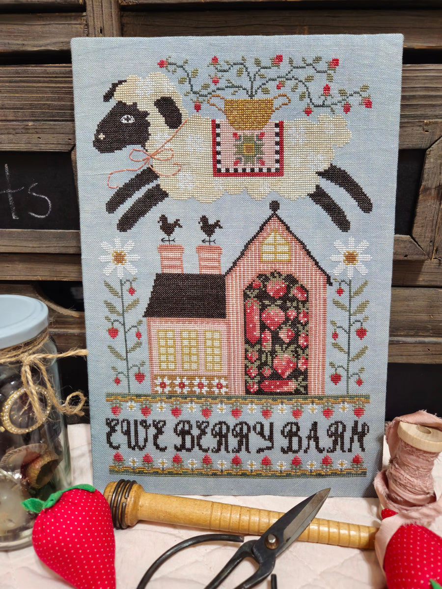 Cross-stitched design of a sheep, house, and text 'Two Bears Barn' on fabric with sewing tools.