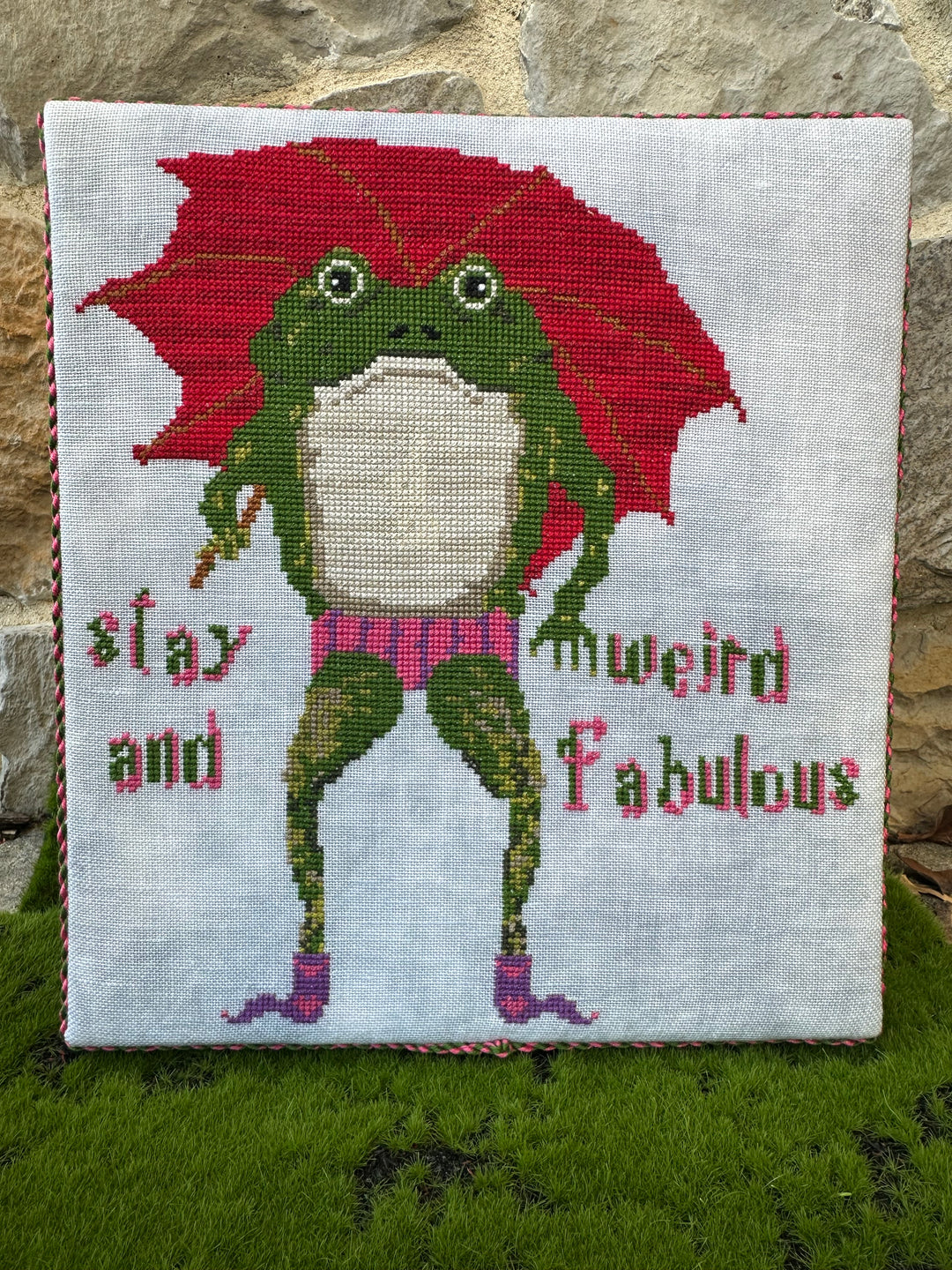 Cross-stitched fabric with a green fairy holding a red umbrella and text on a stone surface.
