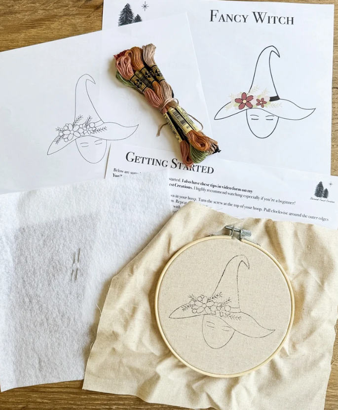 Embroidered witch hat pattern kit on a wooden table, featuring printed pattern sheets with a witch hat design, a bundle of embroidery floss, a wooden embroidery hoop with partially finished beige fabric design, a white felt pad, and a Getting Started instruction sheet.