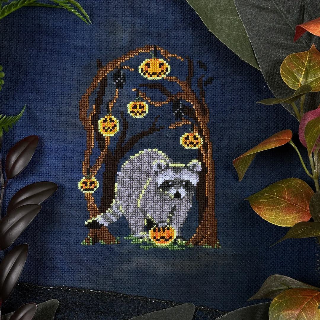 Cross-stitched design of a raccoon with pumpkins on dark fabric, surrounded by leaves.
