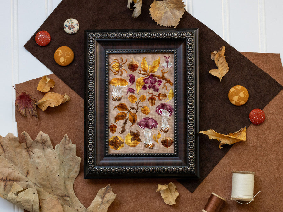 Framed cross-stitched artwork of mushrooms on a brown surface with decorative elements.