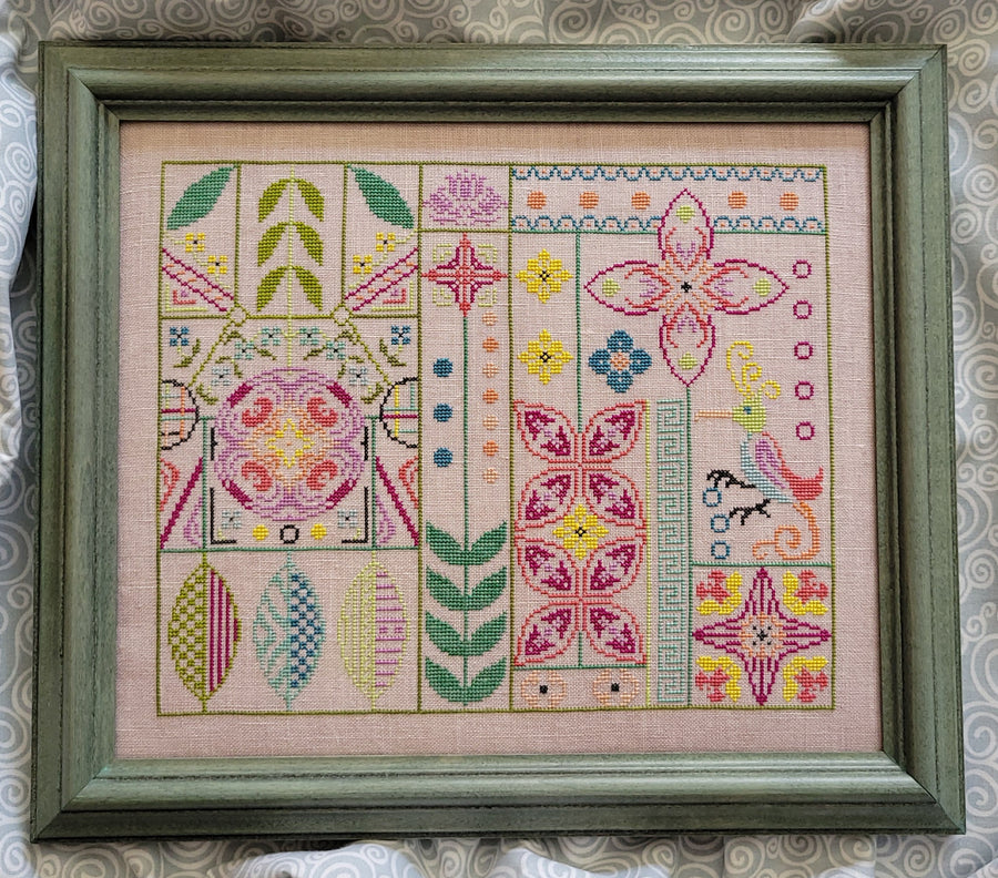Framed cross-stitched pattern with colorful geometric designs on a pink background.