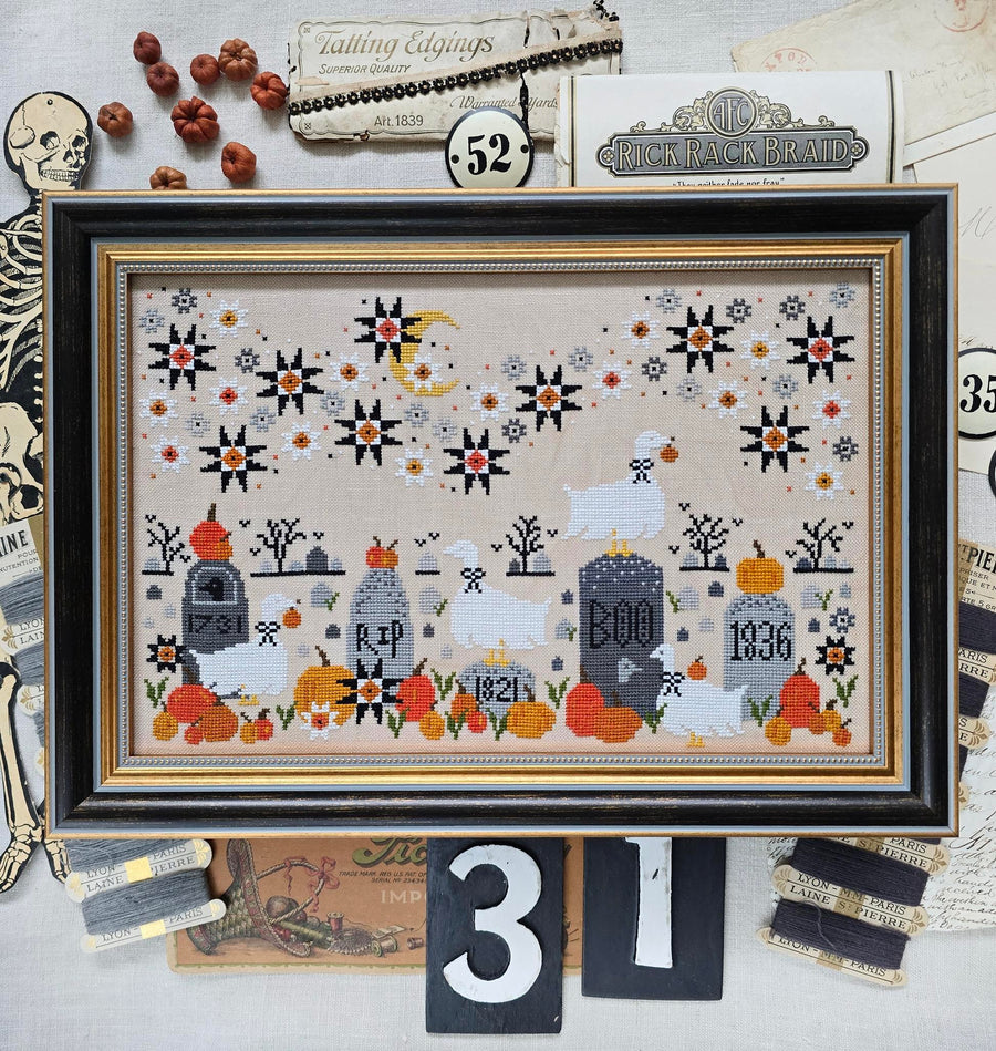 A framed cross-stitch artwork featuring a Halloween cemetery scene with tombstones, ghosts, pumpkins, stars, and a crescent moon, displayed on a styled flat-lay with vintage paper and decorative props.