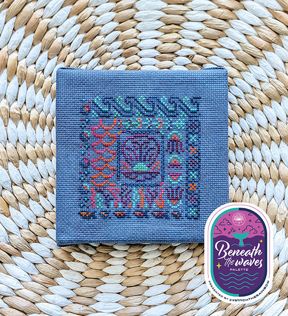 Cross stitch pattern on fabric with 'Beneath the Waves' logo on a woven background