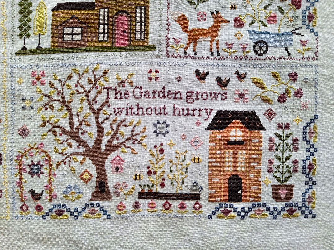 A close-up of a cross-stitch textile featuring a garden-themed design with the phrase 'The Garden grows without hurry' stitched in the center. The design includes a large tree, a small house, flowers, birds, a fox, a wheelbarrow, and decorative motifs on an off-white fabric background.