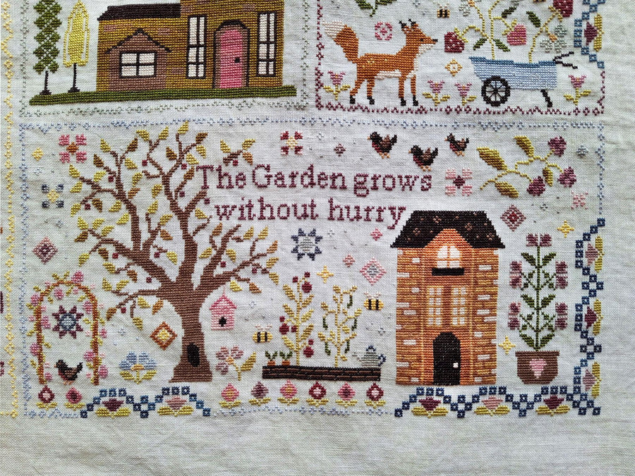 A close-up of a cross-stitch textile featuring a garden-themed design with the phrase 'The Garden grows without hurry' stitched in the center. The design includes a large tree, a small house, flowers, birds, a fox, a wheelbarrow, and decorative motifs on an off-white fabric background.